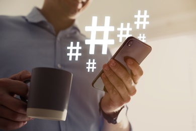Man with cup of drink using modern smartphone, closeup. Hashtag symbols over device