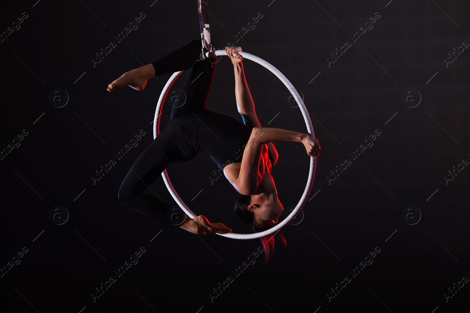 Photo of Young woman performing acrobatic element on aerial ring against dark background