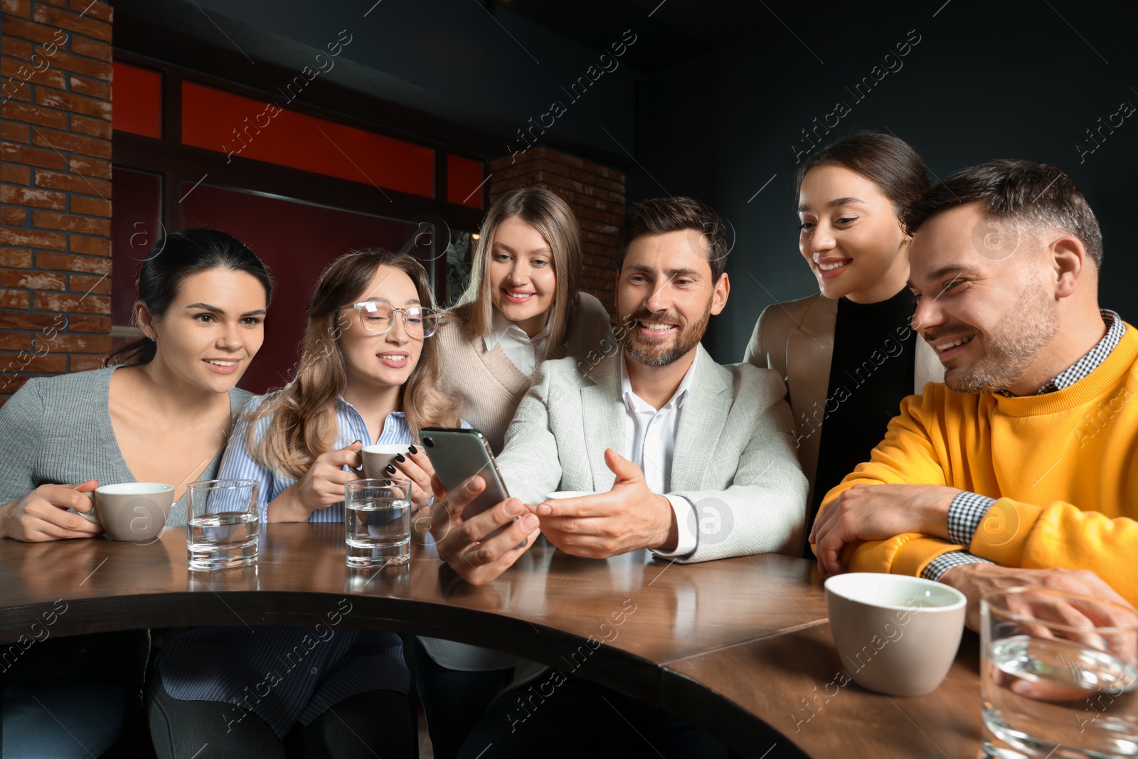 Photo of Handsome man showing something funny in smartphone to his friends in cafe