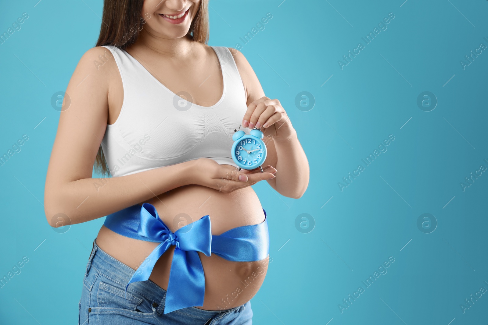 Photo of Young pregnant woman with alarm clock and bow on light blue background, closeup. Time to give birth