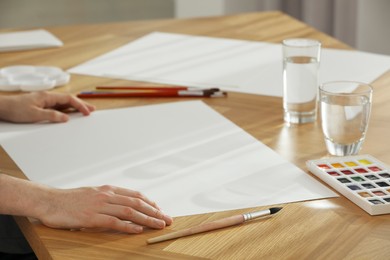 Photo of Man with brushes, paint and blank papers at wooden table indoors, closeup. Watercolor artwork
