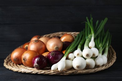 Photo of Wicker mat with different kinds of onions on black wooden table, closeup