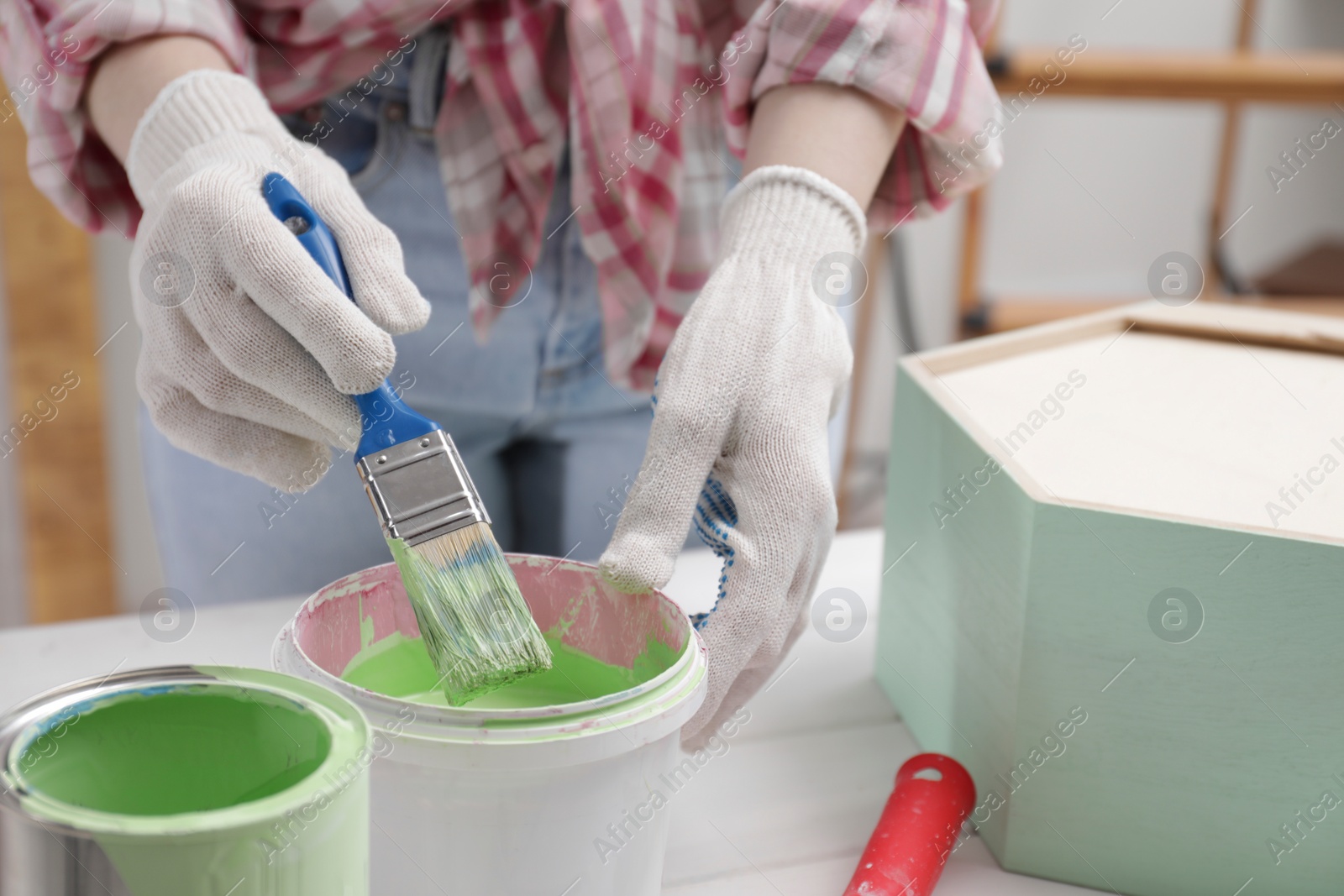 Photo of Woman dipping brush into bucket of green paint at white table indoors, closeup