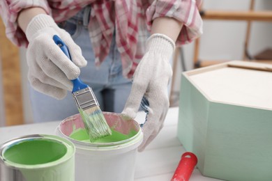 Woman dipping brush into bucket of green paint at white table indoors, closeup