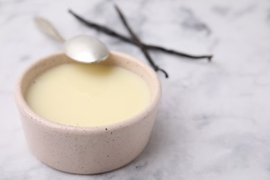 Photo of Bowl with condensed milk, vanilla pods and spoon on white marble table, closeup. Space for text