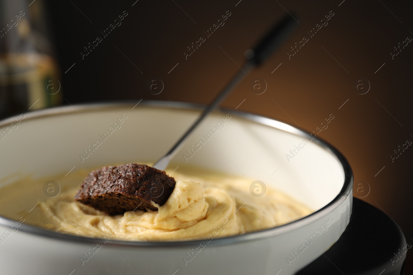 Photo of Fork with piece of bread and melted cheese in fondue pot on dark background, closeup