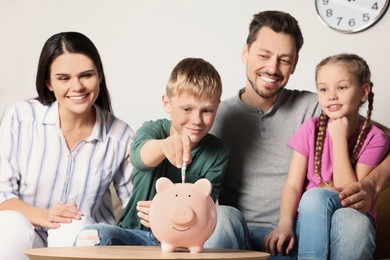 Photo of Happy family putting money into piggy bank at table in room