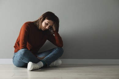 Photo of Sad young woman sitting on floor near grey wall indoors, space for text