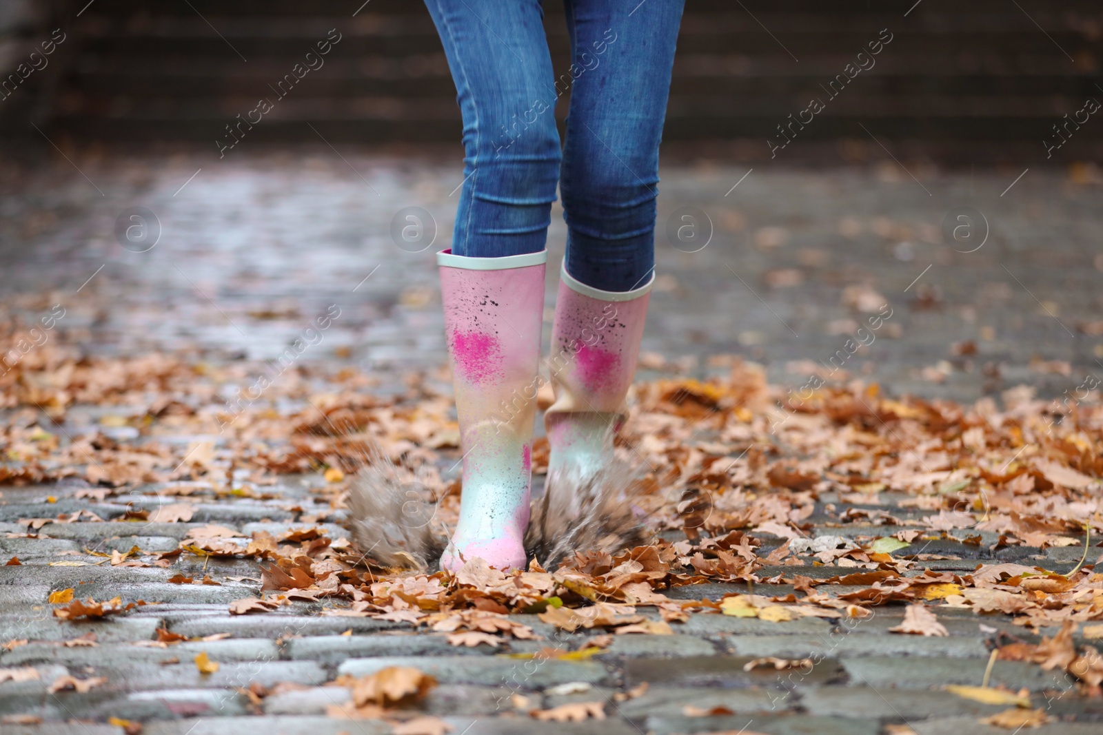 Photo of Woman wearing rubber boots splashing in puddle after rain, focus on legs. Autumn walk