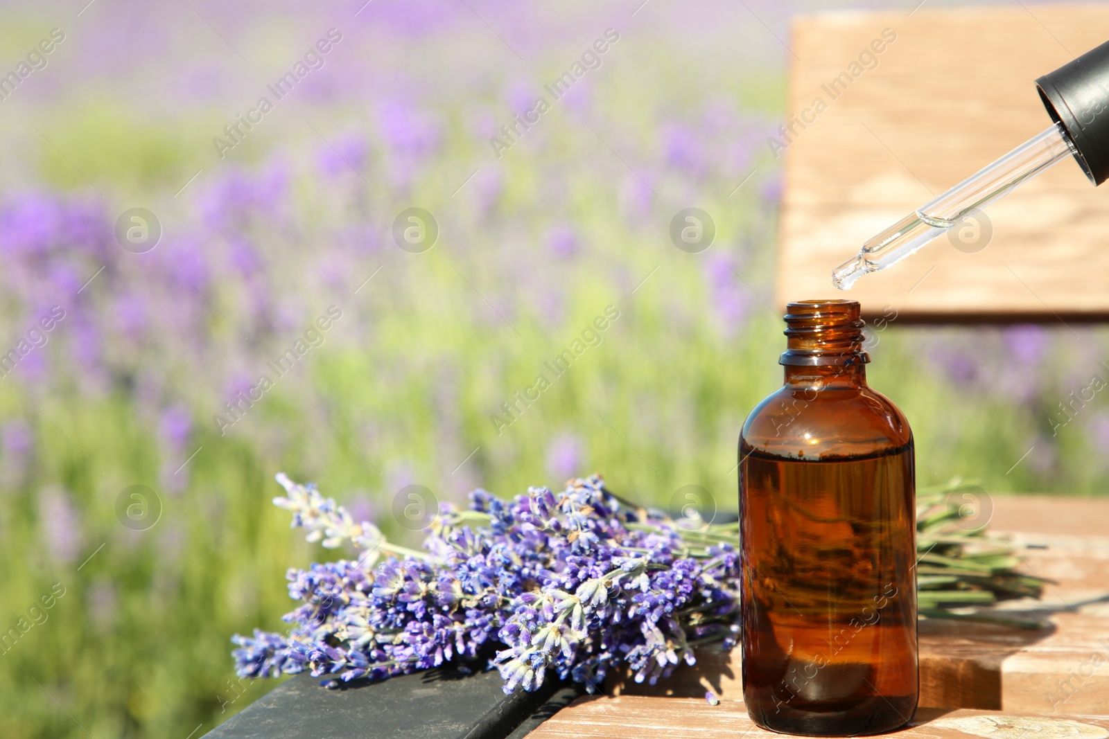 Photo of Dripping essential oil from pipette into bottle near lavender on wooden table outdoors, closeup. Space for text