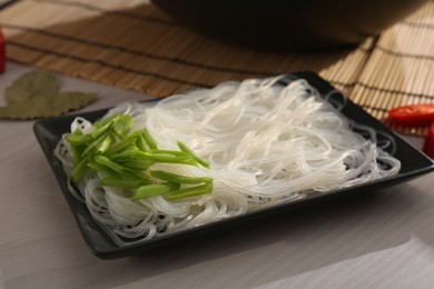 Photo of Plate with rice noodles and green onions near wok on white wooden table, closeup