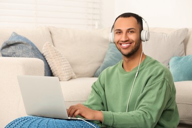 Photo of Portrait of smiling African American man in headphones with laptop near sofa at home