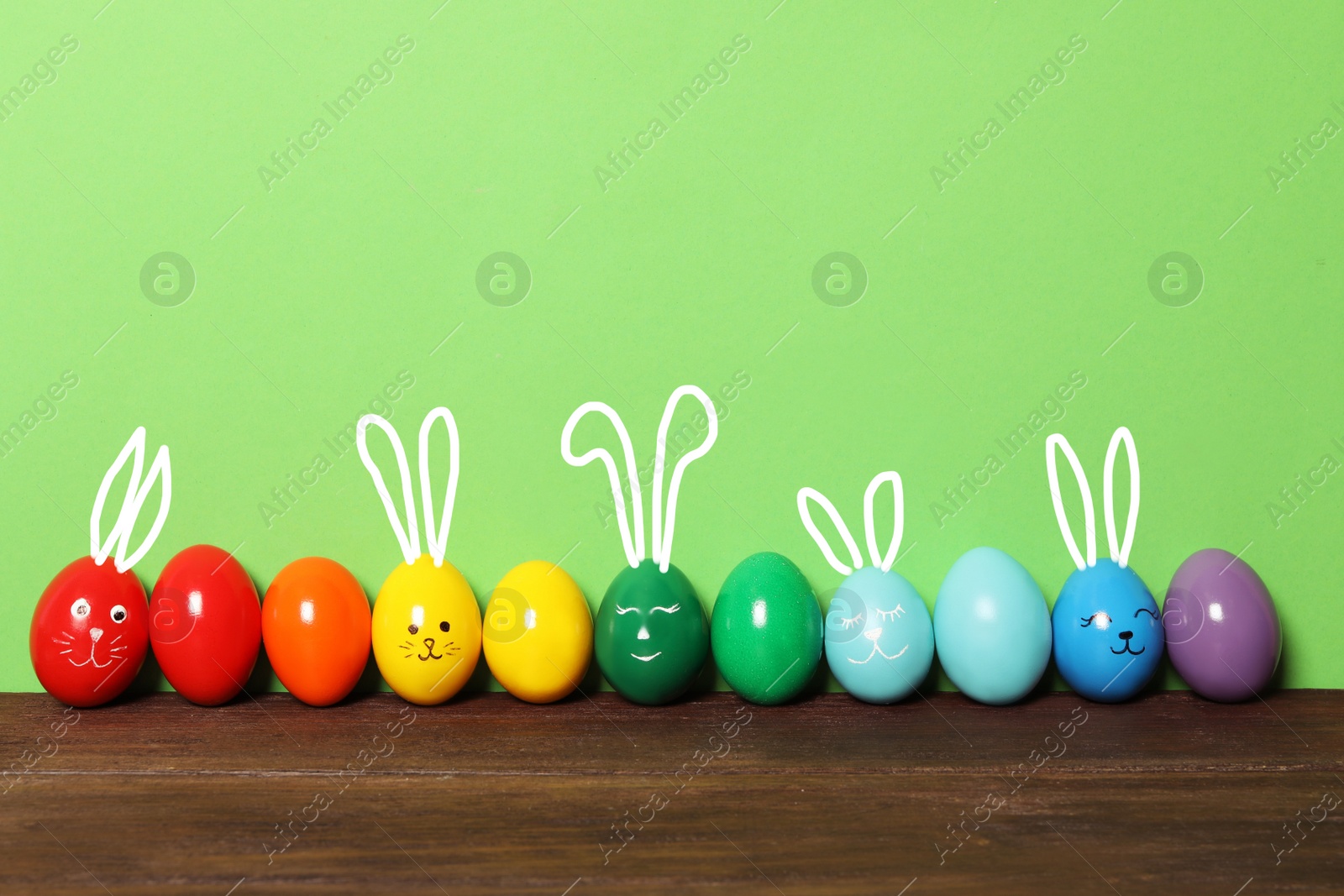 Image of Several eggs with drawn faces and ears as Easter bunnies among others  on wooden table against green background