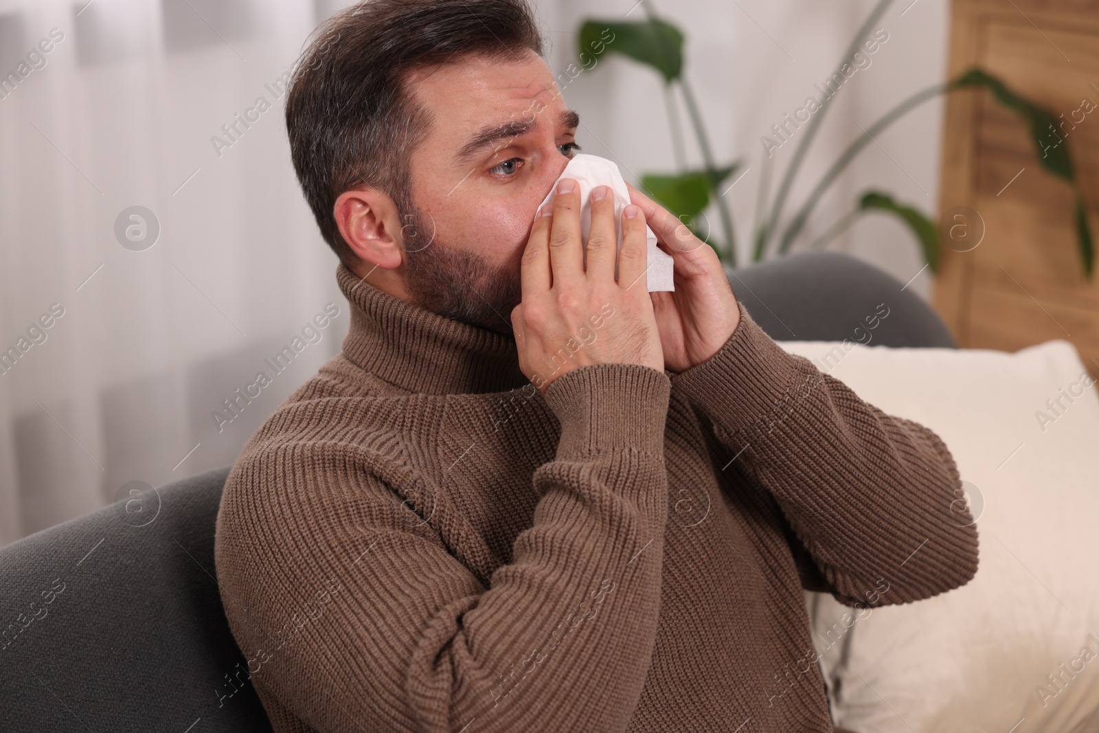 Photo of Sick man with tissue blowing nose on sofa at home. Cold symptoms