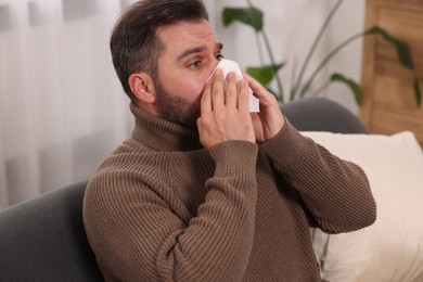 Photo of Sick man with tissue blowing nose on sofa at home. Cold symptoms