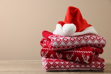 Stack of different Christmas sweaters and Santa hat on wooden table against beige background, space for text