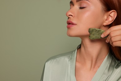 Photo of Young woman massaging her face with jade gua sha tool on green background, closeup. Space for text