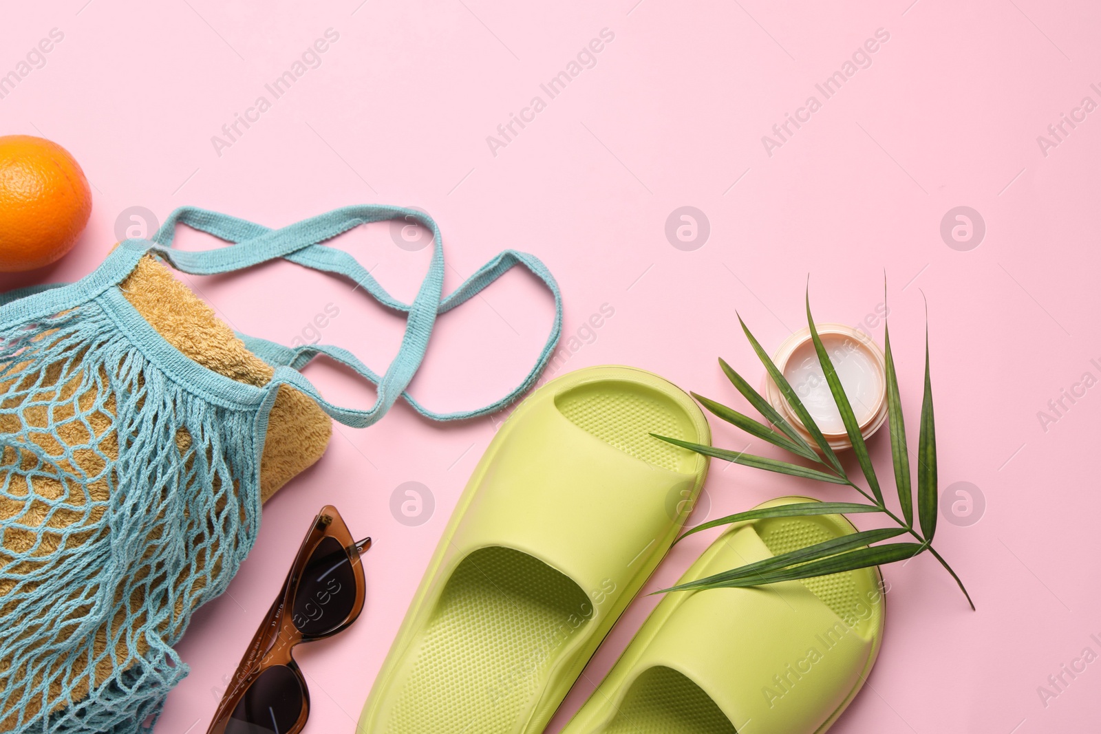 Photo of String bag with towel, sunglasses, orange, slippers, cream and palm leaf on pink background, flat lay