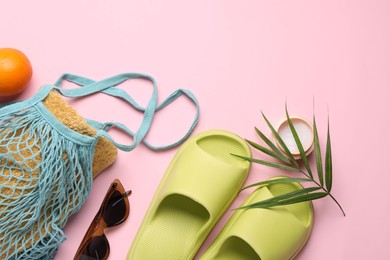 String bag with towel, sunglasses, orange, slippers, cream and palm leaf on pink background, flat lay