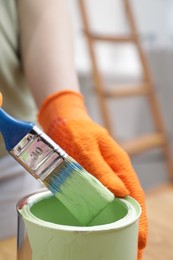 Photo of Woman dipping brush into can of green paint indoors, closeup