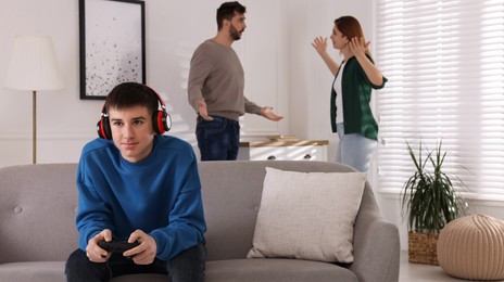 Photo of Teenage boy playing videogame while his parents arguing on background. Problems at home