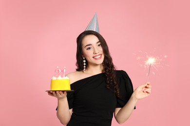 Coming of age party - 21st birthday. Smiling woman holding delicious cake with number shaped candles and sparkler on pink background