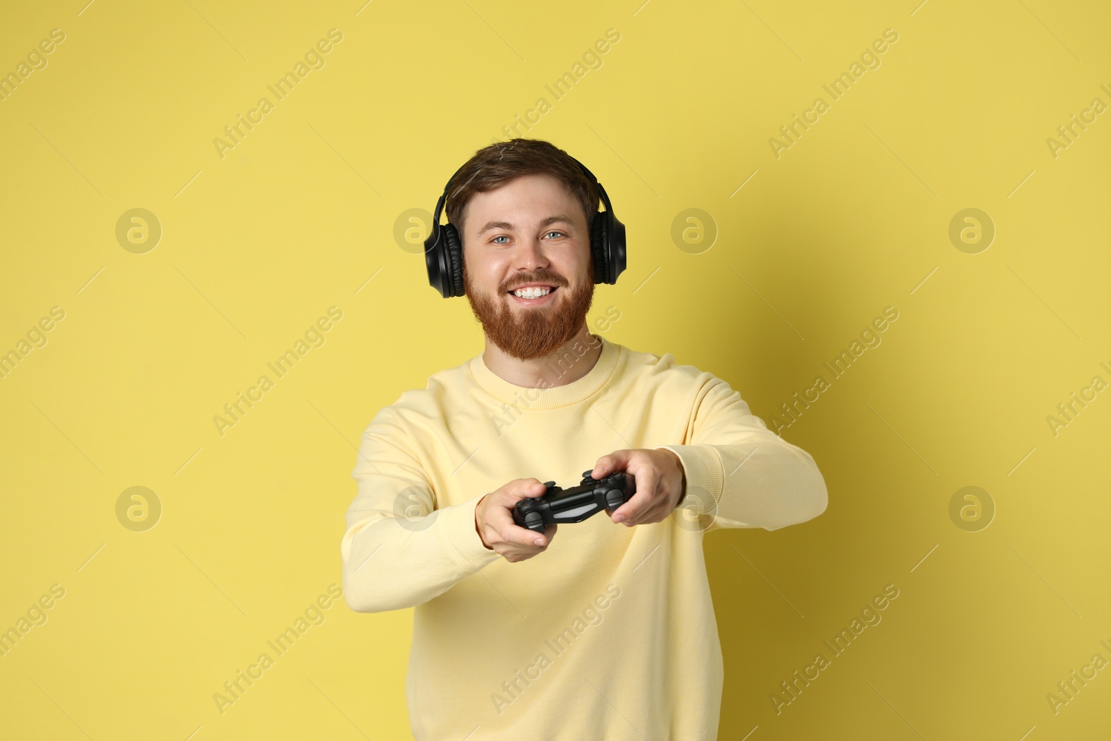 Photo of Happy man in headphones playing video game with controller on pale yellow background