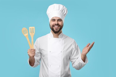 Photo of Happy young chef in uniform holding wooden utensils on light blue background