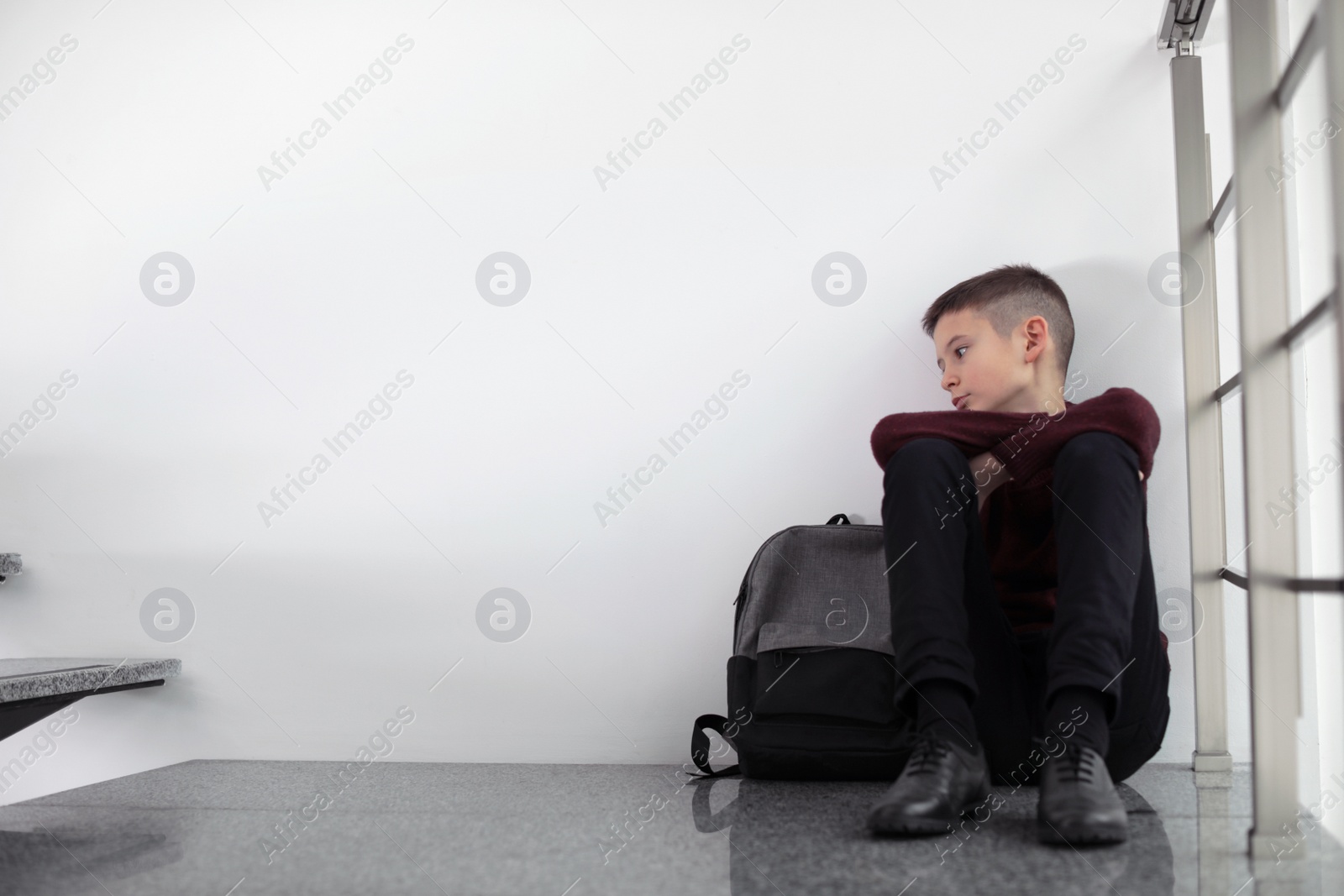 Photo of Upset boy with backpack sitting on staircase indoors. Space for text