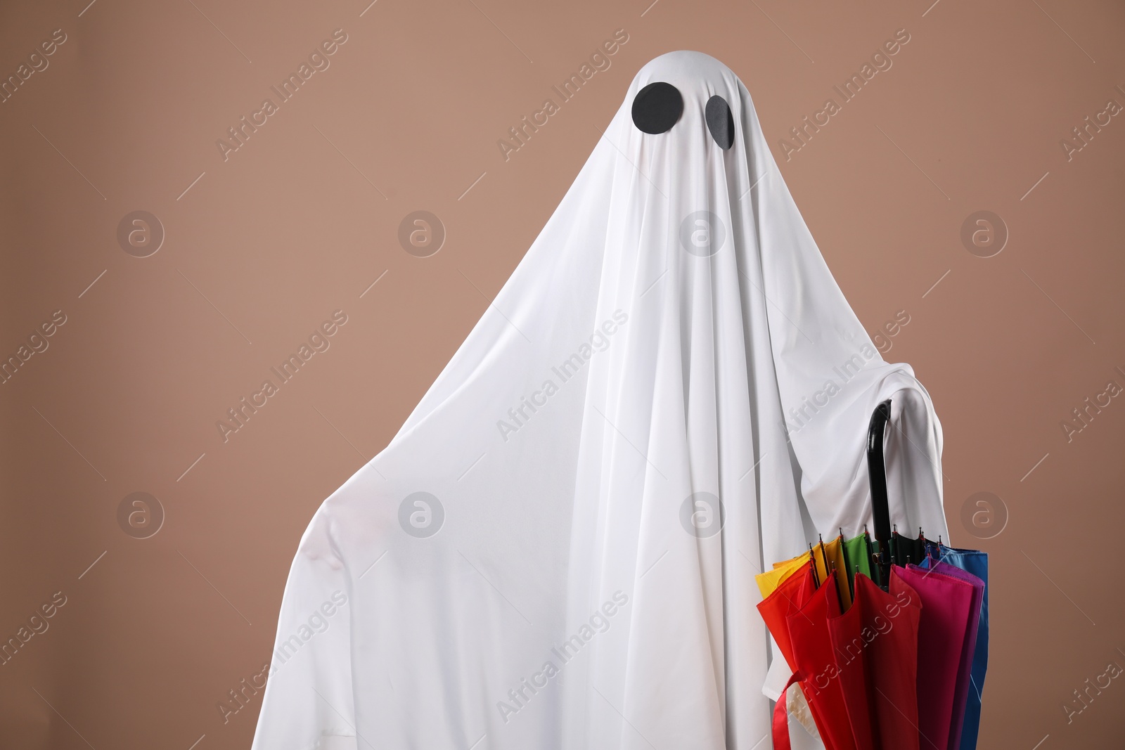 Photo of Person in ghost costume with rainbow umbrella on dark beige background, space for text