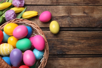 Photo of Colorful Easter eggs in wicker basket and tulips on wooden table, flat lay. Space for text
