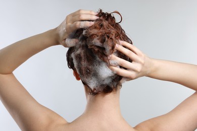 Young woman washing her hair with shampoo on light grey background, back view