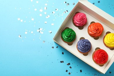 Box with delicious colorful cupcakes and confetti on light blue background, flat lay. Space for text