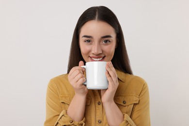 Happy young woman holding white ceramic mug on light grey background