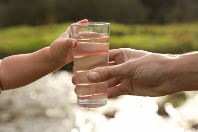 Photo of Father giving her daughter glass of fresh water near stream on sunny day, closeup