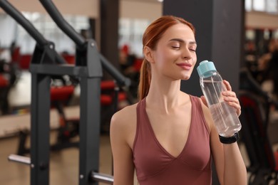 Beautiful young woman with bottle of water in gym, space for text