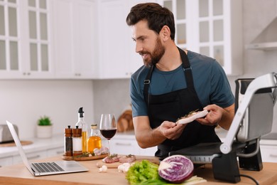 Photo of Man making dinner while watching online cooking course via laptop in kitchen