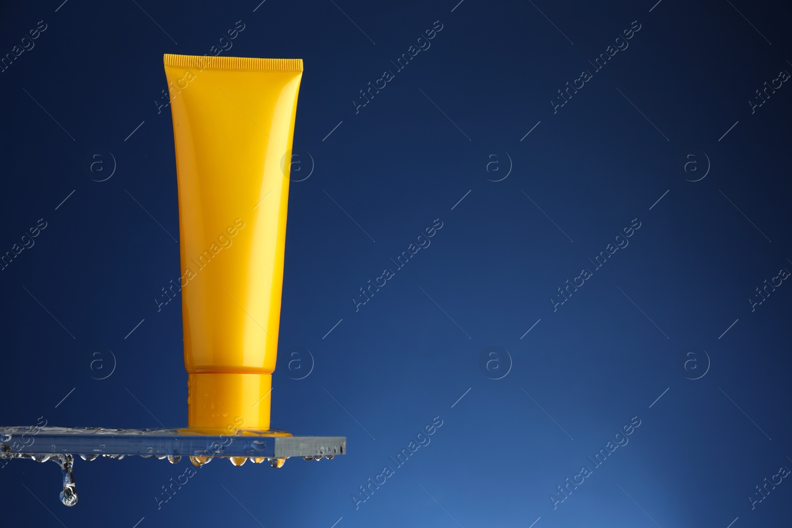 Photo of Moisturizing cream in tube on glass with water drops against blue background. Space for text