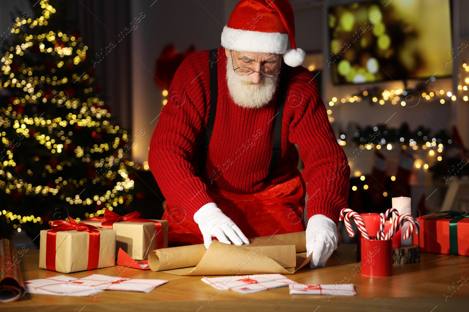 Photo of Santa Claus wrapping gift at his workplace in room decorated for Christmas