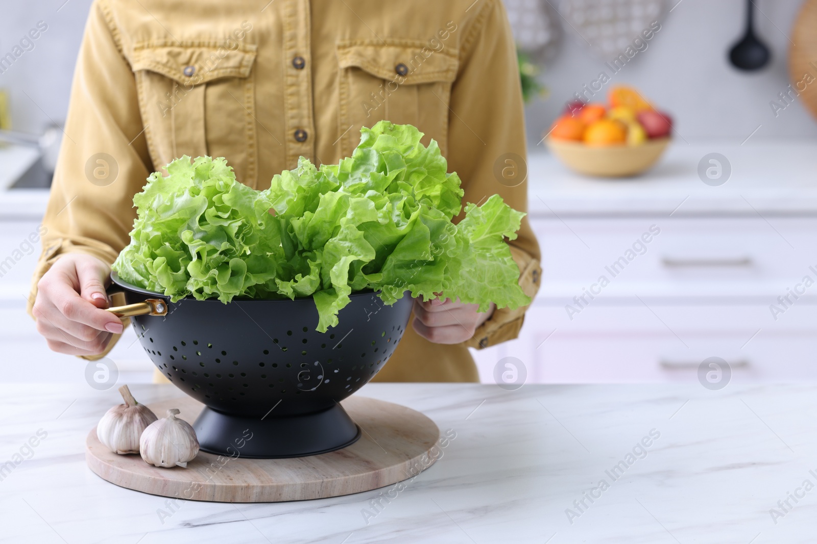 Photo of Woman holding black colander with lettuce at white marble table in kitchen, closeup. Space for text