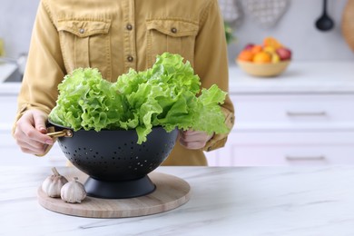 Photo of Woman holding black colander with lettuce at white marble table in kitchen, closeup. Space for text