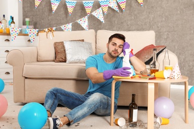 Photo of Tired young man with bottle of detergent in messy room after party