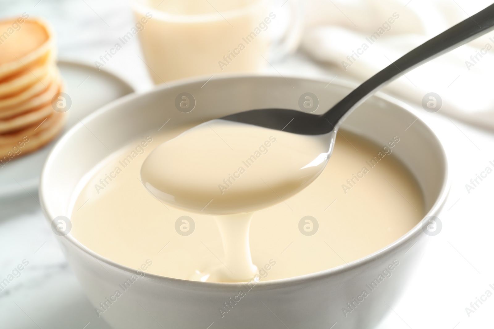 Photo of Spoon of pouring condensed milk over bowl on table, closeup. Dairy products