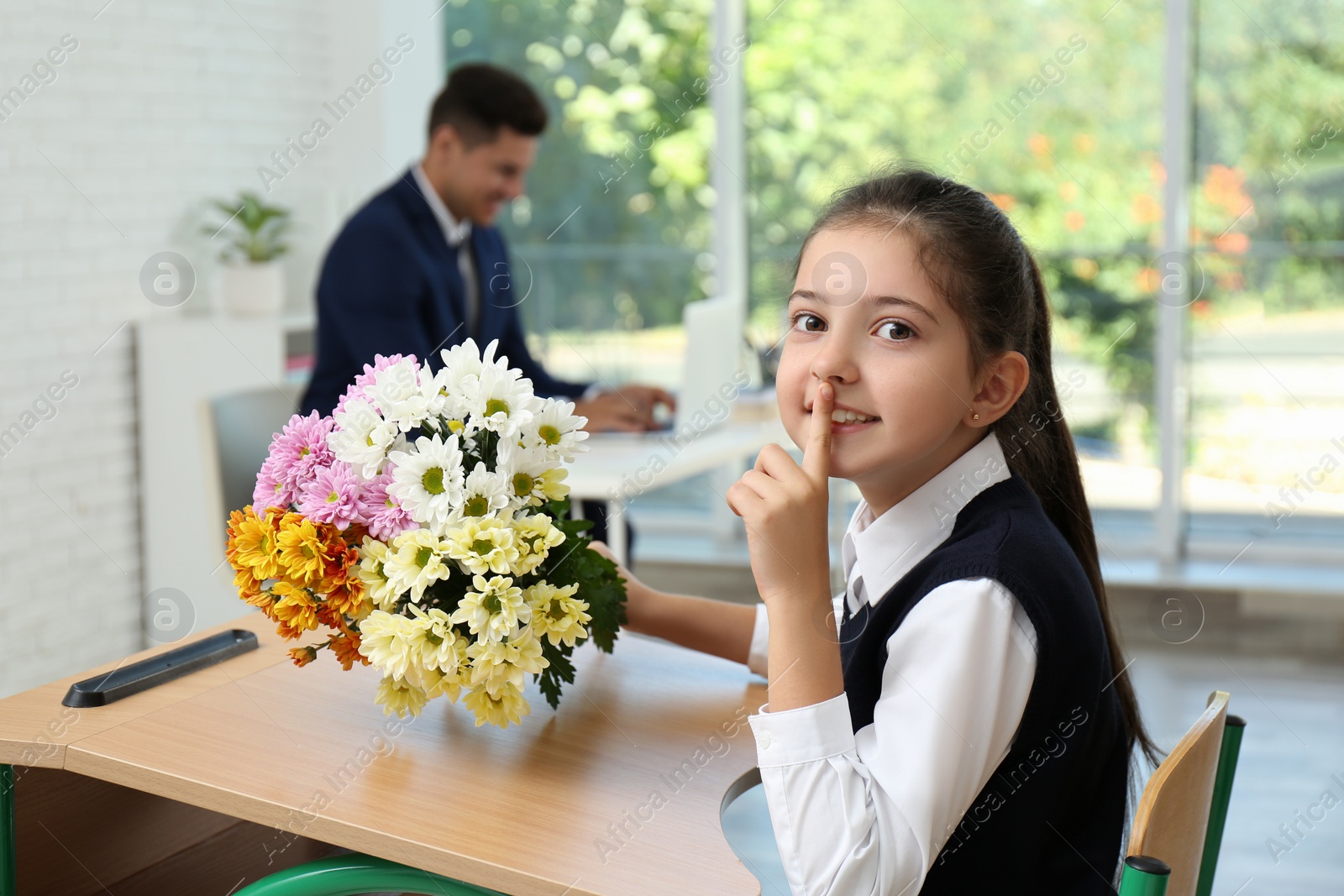 Photo of Happy schoolgirl with bouquet sitting at desk in classroom. Teacher's day