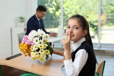 Photo of Happy schoolgirl with bouquet sitting at desk in classroom. Teacher's day
