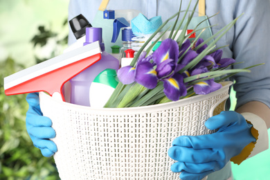 Photo of Woman holding basket with spring flowers and cleaning supplies on green blurred background, closeup