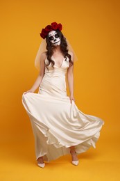 Photo of Young woman in scary bride costume with sugar skull makeup and flower crown posing on orange background. Halloween celebration