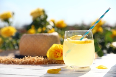 Composition with glass of refreshing lemonade and beautiful rose on white wooden table in blooming garden. Space for text