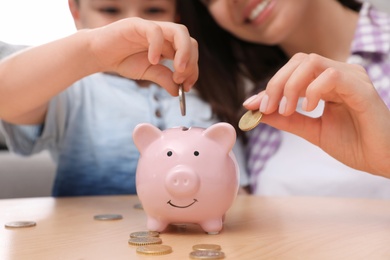 Photo of Happy mother and son putting coins into piggy bank at home, focus on hands