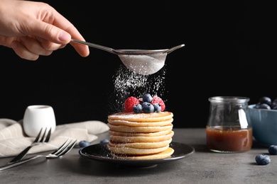 Woman adding sugar powder to tasty pancakes with berries on plate, closeup